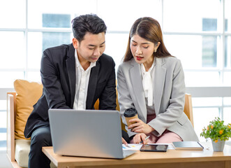 Fototapeta na wymiar Millennial Asian young professional successful businessman employee in formal suit sitting on sofa smiling typing laptop computer while businesswoman colleague holding coffee cup helping advising
