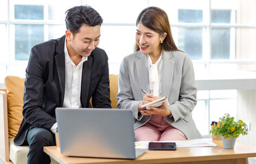 Millennial Asian young professional successful businessman employee in formal suit sitting on sofa smiling typing laptop computer while businesswoman colleague holding coffee cup helping advising