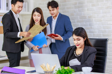 Asian professional successful businesswoman employee sitting working with laptop notebook computer on workstation desk while male and female colleagues standing talking discussing together in office