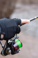 A closeup of a man hand holding a fishing rod. A reel with a green thread can be seen. A blurry background