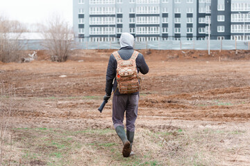 A young man with a hunting backpack is walking near a high building on the cold autumn weather holding a long rod. Back view