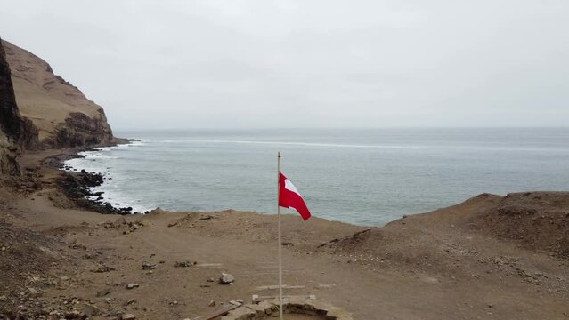 Drone video of a Peruvian flag post near a cliff edge on ocean coast. Drone flies over the flag and forward to the rocky shore. Recorded on winter day in "La Herradura" beach in Chorrillos Lima, Peru.