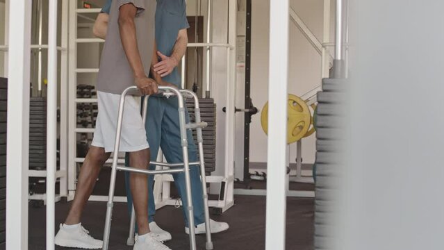 Cropped slowmo of Black man with walkers supported by physiotherapy specialist in blue scrubs walking together along modern gym in rehabilitation center