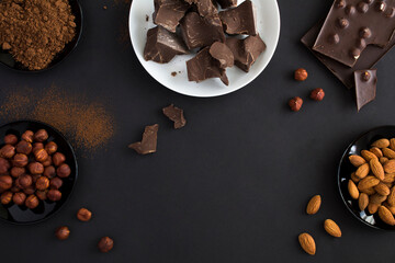 Top view of chunks of dark chocolate on white plate, chocolate, cocoa powder and nuts on the black...
