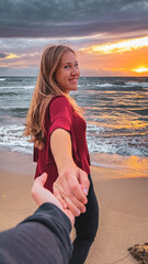 Fototapeta na wymiar Beautiful young blonde woman smiling holding hands with an unrecognizable man in a beautiful sunset on the beach