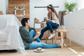 Cute couple eating a sandwich while relaxing from moving in their new house arround cardboard boxes