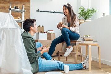 Cute couple eating a sandwich while relaxing from moving in their new house arround cardboard boxes