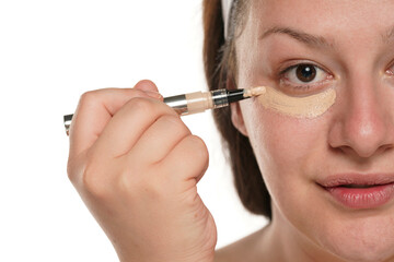 Half portrait of a young smiling chubby woman applyes concealer under her eyes on white background