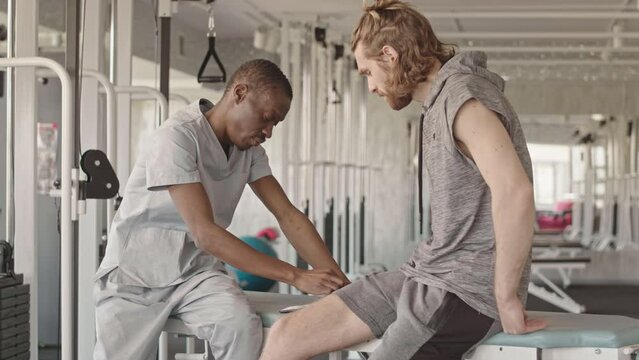 Medium slowmo of African American physiotherapist talking to Caucasian male patient with knee injury having personal physiotherapy session at modern rehabilitation center