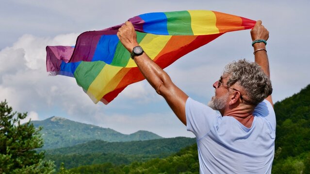 Bisexual, gay, old man, male, transgender hold LGBTQIA flag on sky background, rainbow peace in pride mounts on the nature on a day and celebrate Bisexuality Day or National Coming Out Day