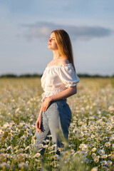 A beautiful redheaded girl is enjoying life in a field of daisies at sunset. A young woman with a wreath of daisies on her head smiles happily in nature.