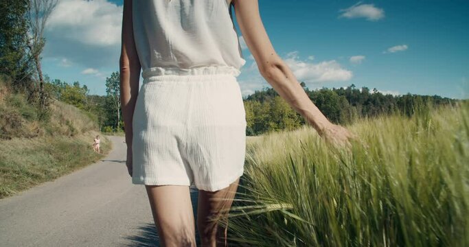 Medium shot of romantic inspired woman walk on country road and touch wheat harvest in slow motion. Children cycling on countryside at background. Summertime vacations on rural environment. 