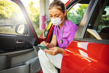 Nurse at car going home and using smartphone. Female doctor in the car wearing a facemask and using...