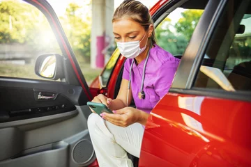 Foto op Canvas Nurse at car going home and using smartphone. Female doctor in the car wearing a facemask and using app on her cell phone - lifestyle concepts. © Jelena Stanojkovic