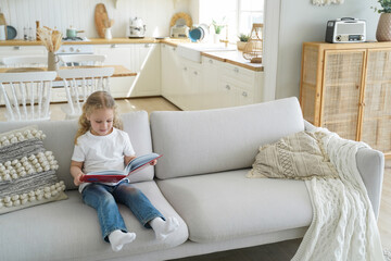 Smart little kid girl reading story holding book sitting on sofa alone at home. Children's education