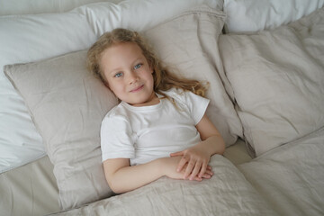 Calm little kid girl covered with duvet lying in bed in after wake up. Healthy sleep in children