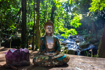 Meditation place, with Buddha statue - Siddhartha Gautama - next to an amethyst stone, in front of a stream, with water running between the rocks in the woods
