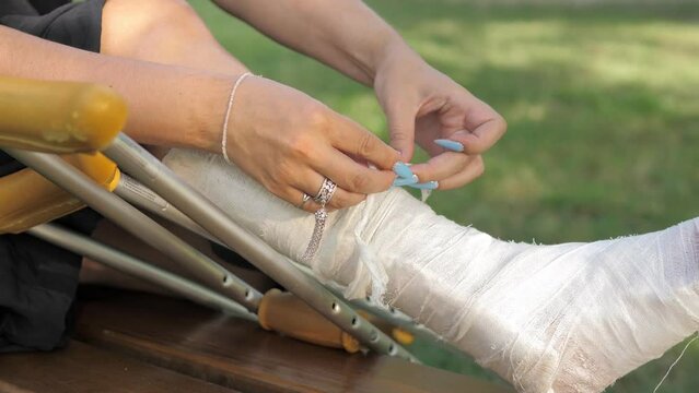 Close-up. A young girl with a cast and a walker is sitting on a bench in the park and tying a knot on her leg bandage.Rehabilitation after a leg strain fracture in the fresh air on a bright sunny day