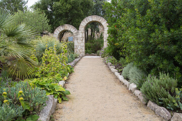 Mediterranean garden or terrace design and landscaping:Beautiful lush green garden with palms, banana trees, foliage plants, perennial herbs along a crushed rock sidewalk leading to stone archways 