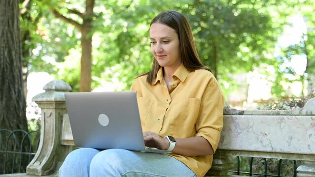 Optimistic young woman using laptop sitting on the bench in campus area, female student watching webinars, educational courses, learning on the distance, typing emails