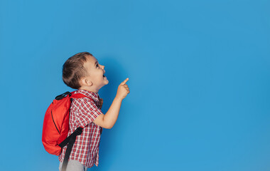 A cheerful smiling little brunette boy with a backpack is having fun on the background of a blue...