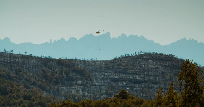 Firefight helicopter is flying over burned mountain forest on terrible wildfire. Ecological catastrophe on Spain natural park at summer with Montserrat view. Fire in Manresa, Pont de Vilomara on July