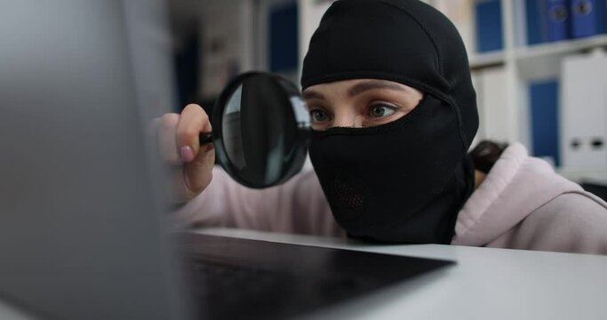 Woman hacker in a balaclava with a magnifier near a laptop