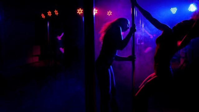 Two sexy adult women dance around a pole in the light of colorful lights in a nightclub. Pole dance.