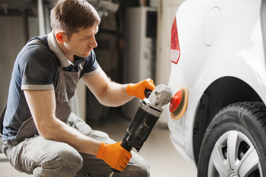 Car detailing - male worker holds a polisher in the hand and polishes white car in auto repair shop.