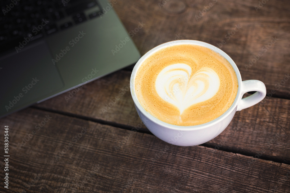 Wall mural Cup of cappuccino with heart next to laptop on wooden table