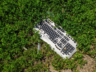 A broken and discarded computer keyboard is buried in the grass.