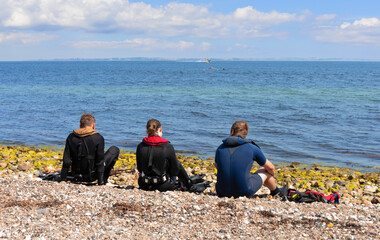 Three people sit on the seashore in diving clothes and look into the distance.