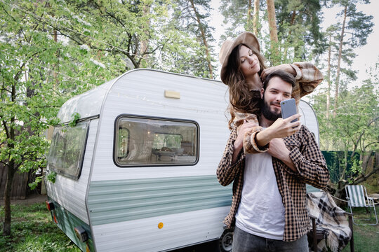 Male giving piggyback ride to girlfriend, having fun and taking a selfie. Happy young caucasian couple traveling in travel van. Romantic atmosphere of relaxation. Road trip around country for weekend