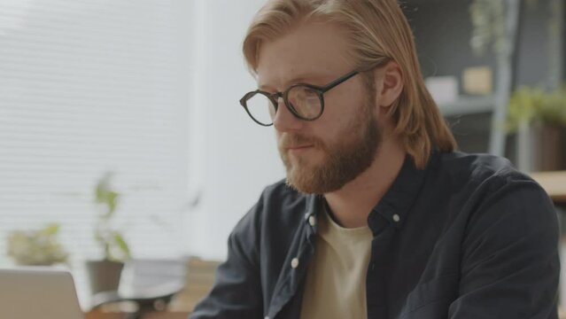 Young bearded businessman in glasses working on laptop and then looking at camera during workday in office
