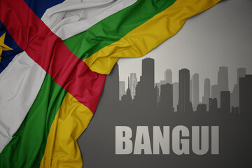 abstract silhouette of the city with text Bangui near waving colorful national flag of central african republic on a gray background.