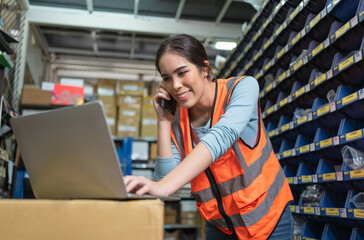 Smiling Asian young female standing and holding smartphone talking with client at warehouse factory.Woman worker using laptop check product order shipment for customer.