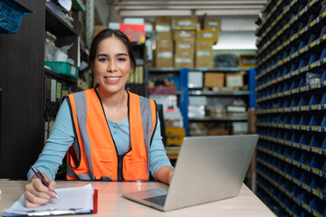 Fototapeta premium Smiling Asian young female sitting and using laptop at warehouse factory.Woman worker in vest safety writing order from customer into paper clipboard in store office.