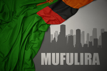 abstract silhouette of the city with text Mufulira near waving colorful national flag of zambia on a gray background.