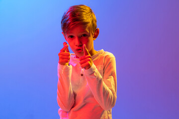 Studio shot of stylish little boy, kid posing isolated on gradient blue-purple background in neon...