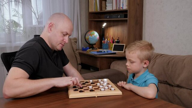 Happy father with son play checkers together at home at the table sitting at the wooden table.