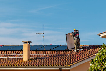 A technician lifts a solar panel to install a photovoltaic system on top of a red-tiled roof