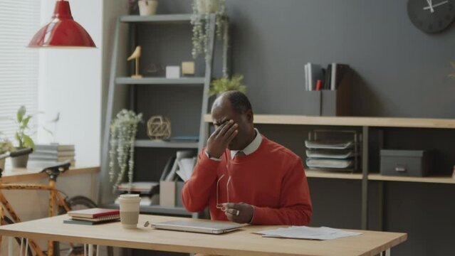 Middle aged African American businessman closing laptop, taking off glasses, rubbing his eyes and leaving office after workday