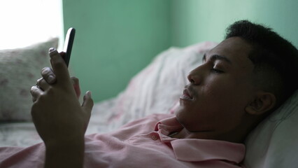 A young hispanic man looking at cellphone device in bedroom lying in bed