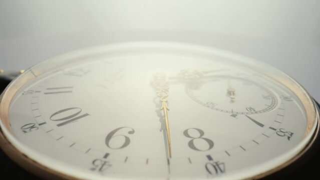 Antique pocket watch with a white dial and gold moving hands is spinning in bright light. The face of an old clock with numbers extra close up. Round mechanical vintage pocket watch.