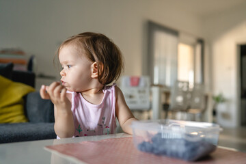 Fototapeta na wymiar One girl child toddler small caucasian female daughter one year old sitting at the table in summer day at home eating blueberries fruit from bowl alone childhood growing up concept real people