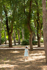 A cute little girl in a summer light dress walks in a park with large trees. The sun. Vertical