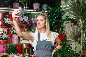 Attractive young florist woman taking a selfie in her flower shop to share on social media posing for the camera on her mobile with a smile. Self employed.