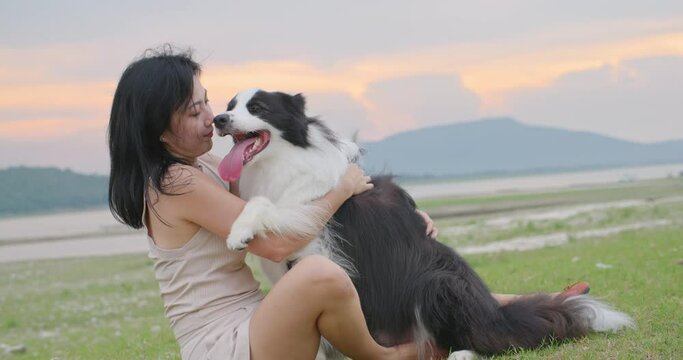 Close up attractive young asian woman hugging and kissing her cute friend black and white border collie dog with love at park on sunset background. Pet lover, Animals and human friendship. Slow motion