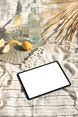 Mockup of new version tablet in trendy thin frame design with white screen lying on a picnic blanket served with a bottle of water and lemons at noon for presentation web design, social media design