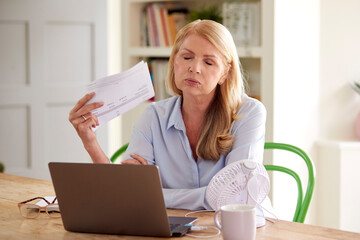 Menopausal Mature Woman Having Hot Flush At Home Cooling Herself With Fan Connected To Laptop